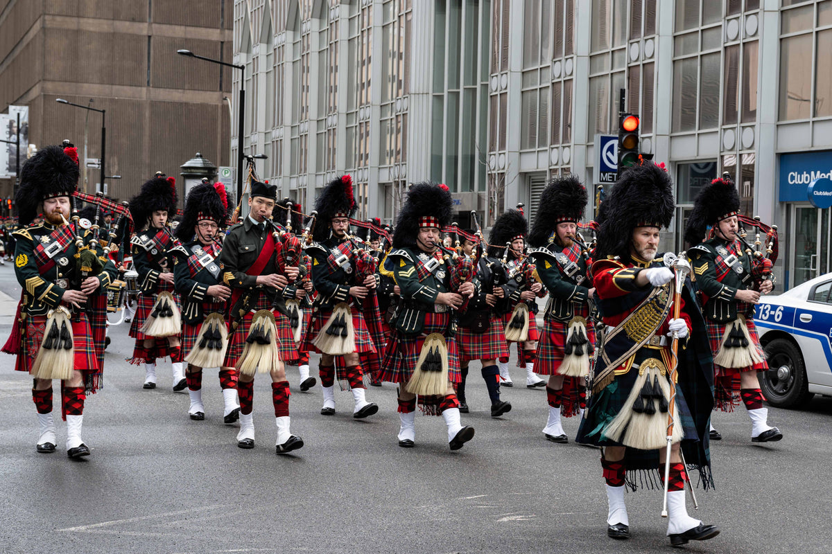 A group of Scottish bagpipers in traditional kilts and tall black bearskin hats marching down a city street, with buildings and a police car visible in the background.