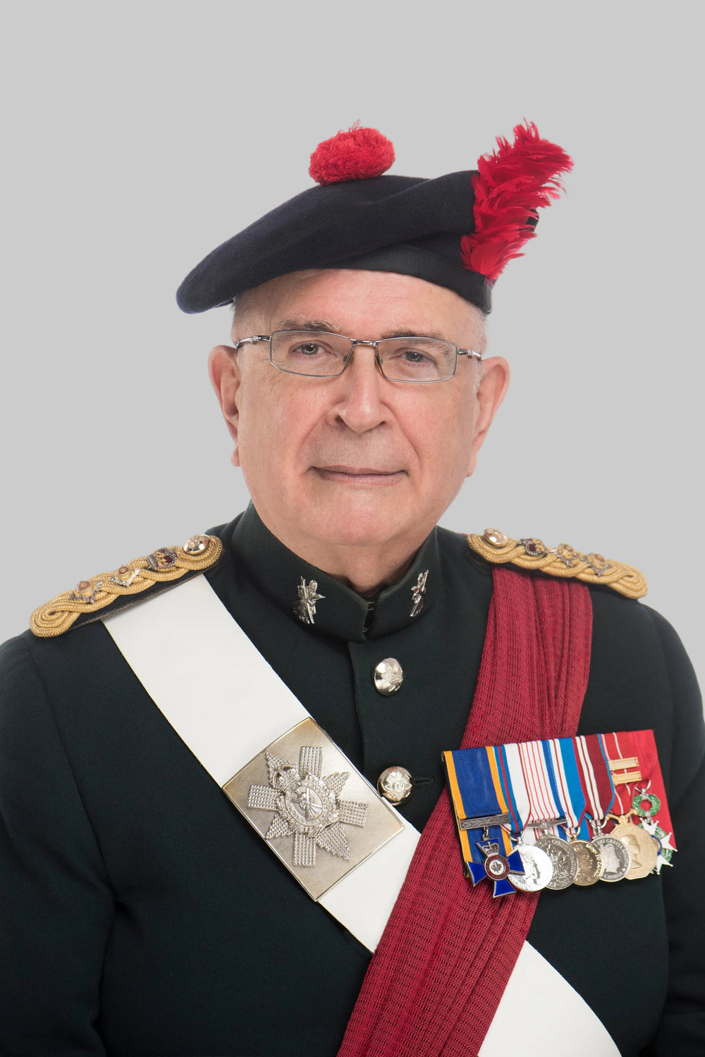 A portrait of a man wearing a black military beret with red pom-poms and a feather, a black uniform jacket with gold epaulettes, a white sash, and multiple medals on the chest, against a plain white background.