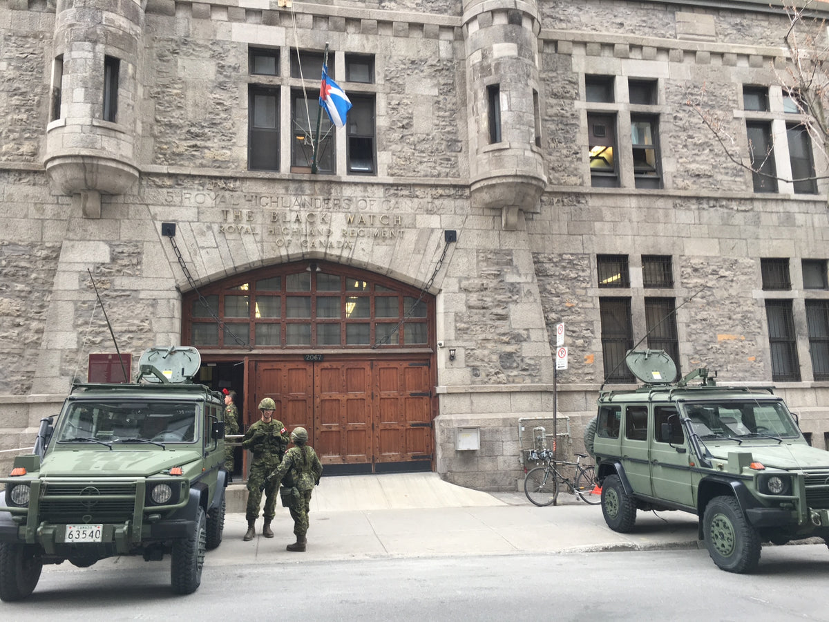 Two military armored vehicles with soldiers standing in front of a large stone building with an arched wooden entrance and a flag above.
