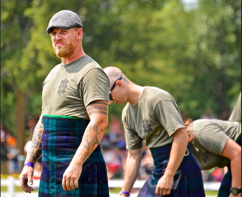 A man outdoors wearing an olive green t-shirt and a blue and purple tartan kilt, with a gray flat cap. Another man in a matching outfit is bent over in the background, with trees and a crowd blurred in the distance.