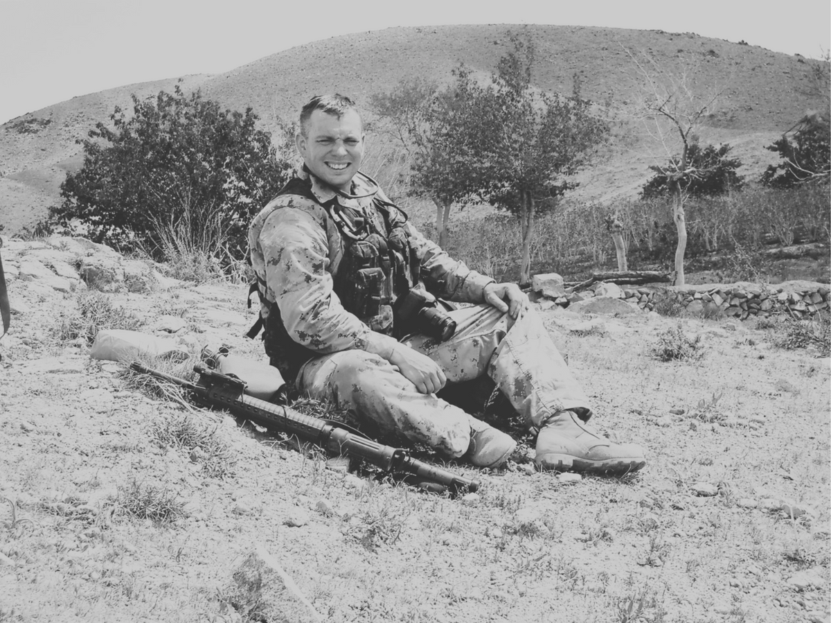 A black and white photograph of a smiling man in military camouflage sitting outdoors on a rocky, grassy hillside with a rifle resting beside him.